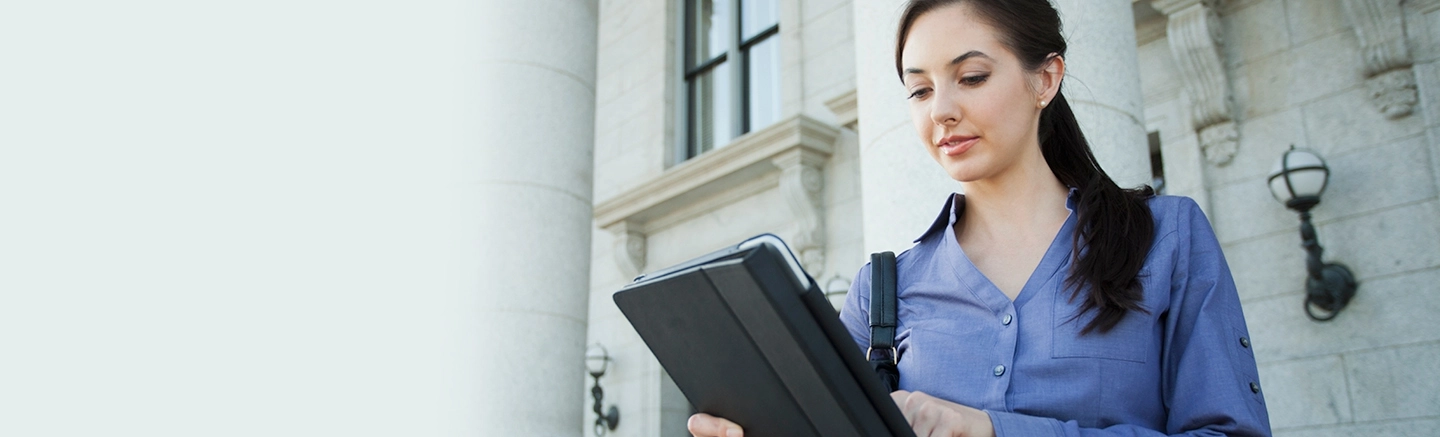 Person in a blue shirt holding a tablet outside a stone building with columns, illustrating how to secure a job interview and effective tactics for job applications.